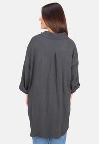 Gray, textured long-sleeve shirt with a loose fit, rollable sleeves, and back pleat detail. Worn with blue jeans.