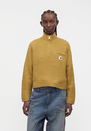 Donna con capelli intrecciati che indossa un maglione a maglia giallo senape con mezza cerniera e jeans larghi azzurri, in piedi contro uno sfondo semplice.