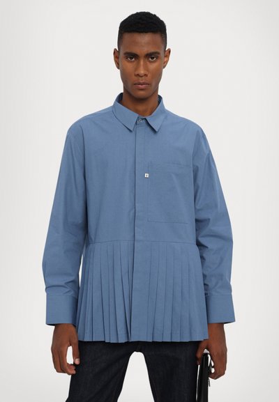 Chemise bleu clair avec un col, des manches longues, une poche poitrine et une partie inférieure plissée. Fabriquée en tissu lisse.