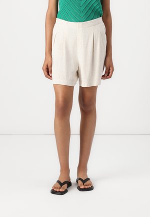 Vrouw draagt beige geplooide korte broek, groen mouwloos gebreid topje en zwarte sandalen met vierkante neus, staand tegen een witte achtergrond.