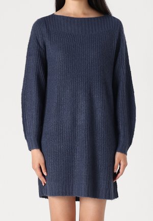 Kobieta ubrana w granatową, strukturalną dzianinową sukienkę z długim rękawem, stojąca z rękami swobodnie wzdłuż ciała.