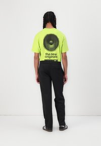 Neon žalia trumpa rankovė marškinėliai su dideliu juodu garsiakalbio grafiku ir tekstu ant nugaros, derinami su juodomis kelnėmis ir juodais batais.