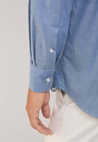 Blauwe chambray overhemdmouw met een puntige manchetknoop, met twee witte knopen en een subtiele structuur; hand gepositioneerd nabij de zoom.