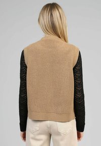 Gilets en tricot beige avec une texture côtelée, présentant un col ras du cou et un design sans manches, porté par-dessus un haut en dentelle noir à manches longues.