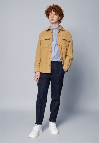 Giacca in corduroy beige con due tasche sul petto, camicia a righe blu e bianche, pantaloni a righe blu navy, e sneakers bianche con dettagli logo.