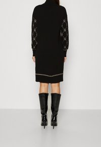 Kvinna i en svart knä-lång klänning med mönstrade, långa ärmar och svarta högklackade knäboots, som står mot en enfärgad vägg.