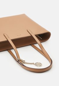 Torebka tote w kolorze tan z długimi paskami oraz złotym charms'em w postaci łańcuszka z wygrawerowanym okrągłym medalionem na białym tle.