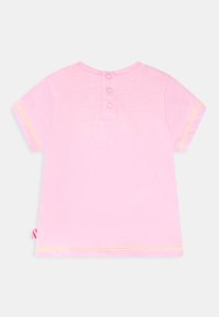 T-shirt de algodão rosa com mangas curtas, fecho traseiro com botão, costura amarela ao longo das mangas e uma pequena etiqueta lateral.