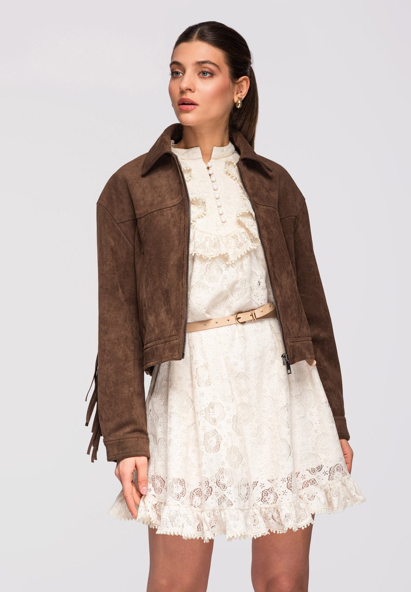 Donna che indossa un vestito bianco in pizzo con balze e bottoni, cintura beige e giacca di suede marrone con maniche frangiate, che guarda leggermente verso destra.