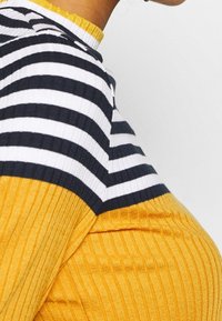 Rippstrickpullover mit einem gestreiften Muster in Marineblau und Weiß im oberen Bereich, und einem unifarbenen unteren Teil in Senfgelb.