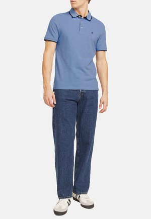 Blauwe polo met zwarte accenten; korte mouwen. Draagt het met losse blauwe jeans en witte sneakers met zwarte strepen.
