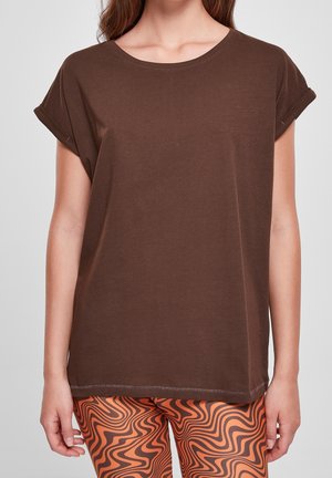 Kobieta ubrana w gładki brązowy t-shirt z krótkim rękawem oraz pomarańczowe legginsy z czarnymi falistymi wzorami, stojąca na szarym tle.