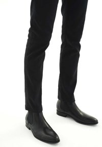 Botas de cuero negras hasta el tobillo con punta afilada, paneles laterales elásticos y un tacón bajo apilado, combinadas con pantalones negros de pierna recta.