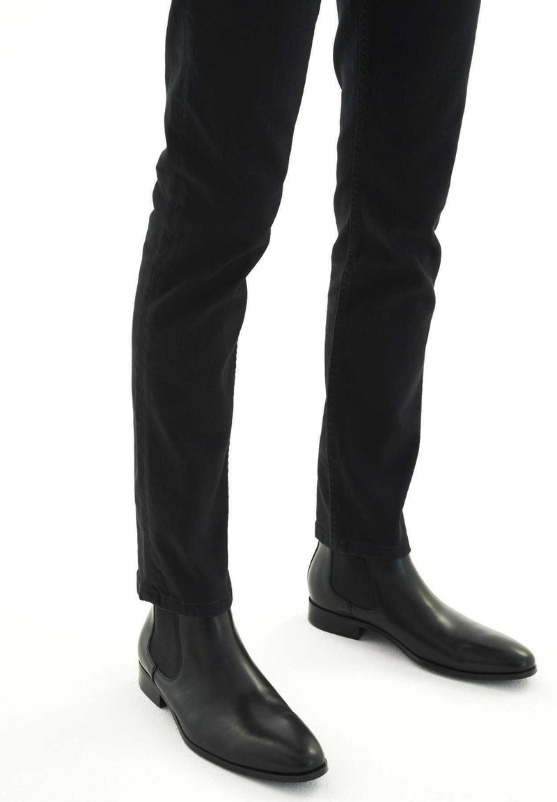 Botas de cuero negras hasta el tobillo con punta afilada, paneles laterales elásticos y un tacón bajo apilado, combinadas con pantalones negros de pierna recta.