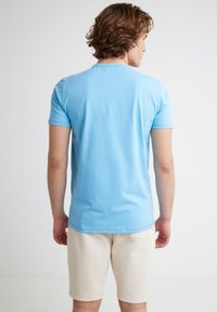 Kreklu ar īsām piedurknēm, gaismas zilā krāsā, izgatavots no kokvilnas, ar apaļo izgriezumu, valkā ar krēmkrāsas šortiem, parādot gludu tekstūru un vienkāršu dizainu.