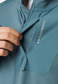 Vaaleansininen vetoketjullinen takki, joka on valmistettu teksturoidusta kankaasta. Siinä on korkea kaulus, sujuva vetoketju ja prominenssi logo rinnassa.