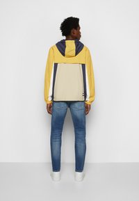 Giacca a vento color block gialla e blu navy con cappuccio, polsini elastici e pannello posteriore beige, abbinata a jeans blu e scarpe da ginnastica bianche.