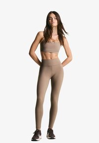 Beige sportstop og højtaljede leggings lavet af glat, strækbart stof. Fodtøjet har mørke accenter. Modellen står med hænderne på hoften.