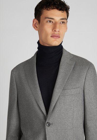 Blazer grigio in lana con un bottone singolo, revers a scendere e tasca sul petto, indossato su un maglione a collo alto blu scuro.