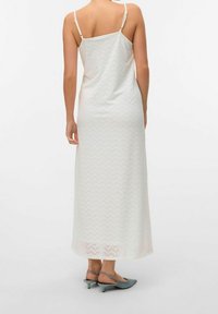 Femme portant une longue robe blanche sans manches avec un motif perforé en chevrons, et des chaussures slingback grises à talons bas, vue de dos.