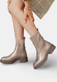 Bottines métalliques dorées avec des côtés élastiques nervurés et une semelle épaisse. Texture lisse avec un zipper à l'arrière.