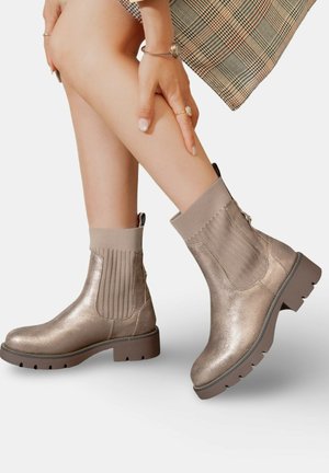 Bottines métalliques dorées avec des côtés élastiques nervurés et une semelle épaisse. Texture lisse avec un zipper à l'arrière.