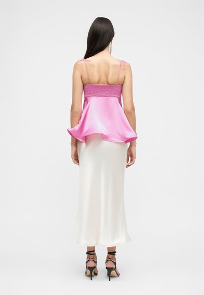 Γυναίκα που φοράει ροζ σατέν μπλουζάκι χωρίς μανίκια με λεπτές τιράντες και άσπρη φούστα μέχρι τον αστράγαλο, εμφανίζεται από πίσω με λευκό φόντο.