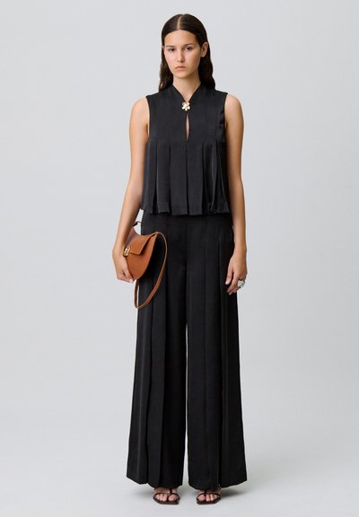 Top negro sin mangas con pliegues y un detalle de ojal, combinado con pantalones anchos negros. Lleva un bolso de hombro marrón, acentuado por un anillo.