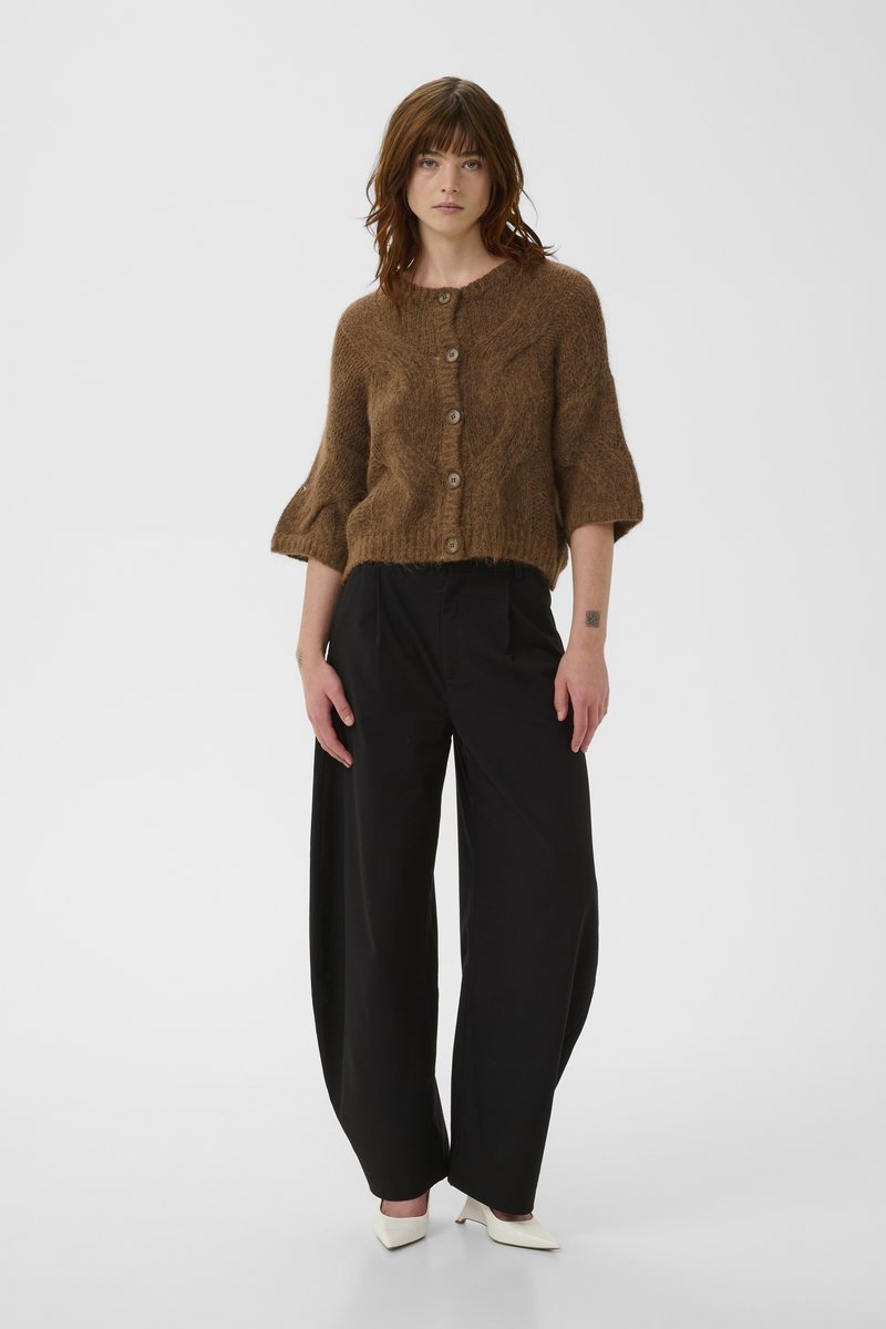 Femme debout portant un cardigan en maille marron boutonné, un pantalon noir à jambes larges et des talons blancs à bouts pointus, sur un fond gris uni.