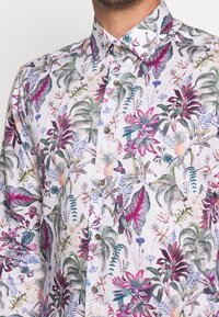 Camicia a maniche corte con sfondo bianco e un vivace motivo floreale tropicale in tonalità di verde, rosa e viola, con bottoni marroni.