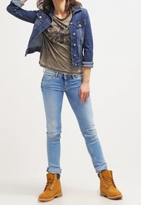 Džínová bunda, olivové tričko s grafikou, světle modré těsné džíny a žluto-hnědé šněrovací boty s texturovaným povrchem a hnědými akcenty.