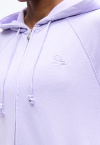 Κοντινό πλάνο ενός ατόμου που φοράει ανοιχτό μοβ φούτερ με φερμουάρ, κουκούλα και κορδόνια, με μικρό λογότυπο Champion στο στήθος.