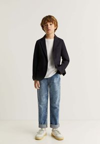 Blazer negro, camiseta blanca, jeans azul desgastados, puños enrollados y zapatillas blancas con suelas beige. Chico de pie contra un fondo liso.