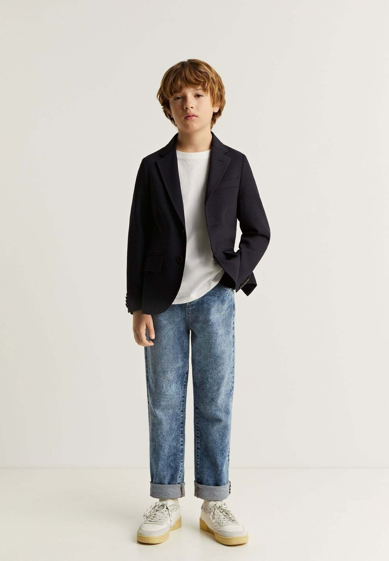 Blazer negro, camiseta blanca, jeans azul desgastados, puños enrollados y zapatillas blancas con suelas beige. Chico de pie contra un fondo liso.