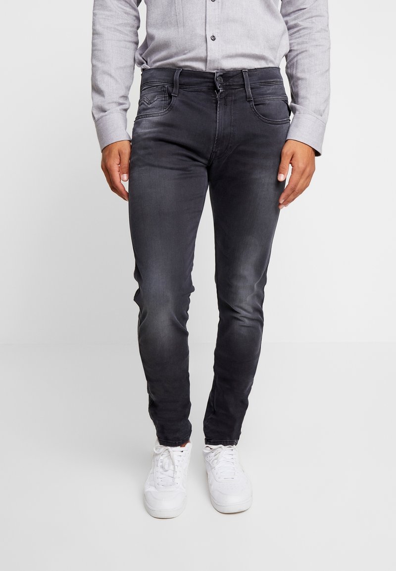 Jean slim gris foncé avec légère décoloration, cinq poches et coutures contrastées. Porté avec une chemise à boutons grise et des baskets blanches.