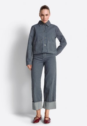 Nő áll, kék-fehér függőlegesen csíkos zakóban és hozzáillő, visszahajtott szegélyű bő szárú nadrágban, piros lapos cipőben.