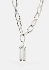 Colar de corrente prateada com elos alongados e um pingente retangular facetado com uma pedra transparente.