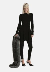 Woman with blonde hair wearing a long-sleeve black dress over black leggings and black heels, holding a large gray fur stole.