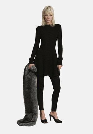 Robe en tricot noire à manches longues, silhouette en A, associée à des leggings noirs et des talons, tenant un long châle en fausse fourrure grise.