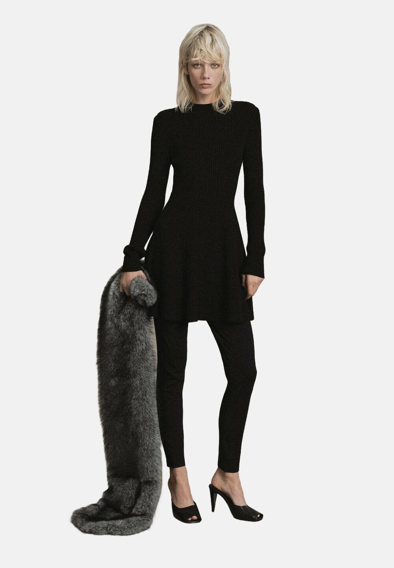 Woman with blonde hair wearing a long-sleeve black dress over black leggings and black heels, holding a large gray fur stole.