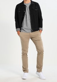 Sort skjortejakke over en grå T-shirt, kombineret med beige bukser og hvide sneakers. Jakken har to brystlommer og op rullede ærmer.