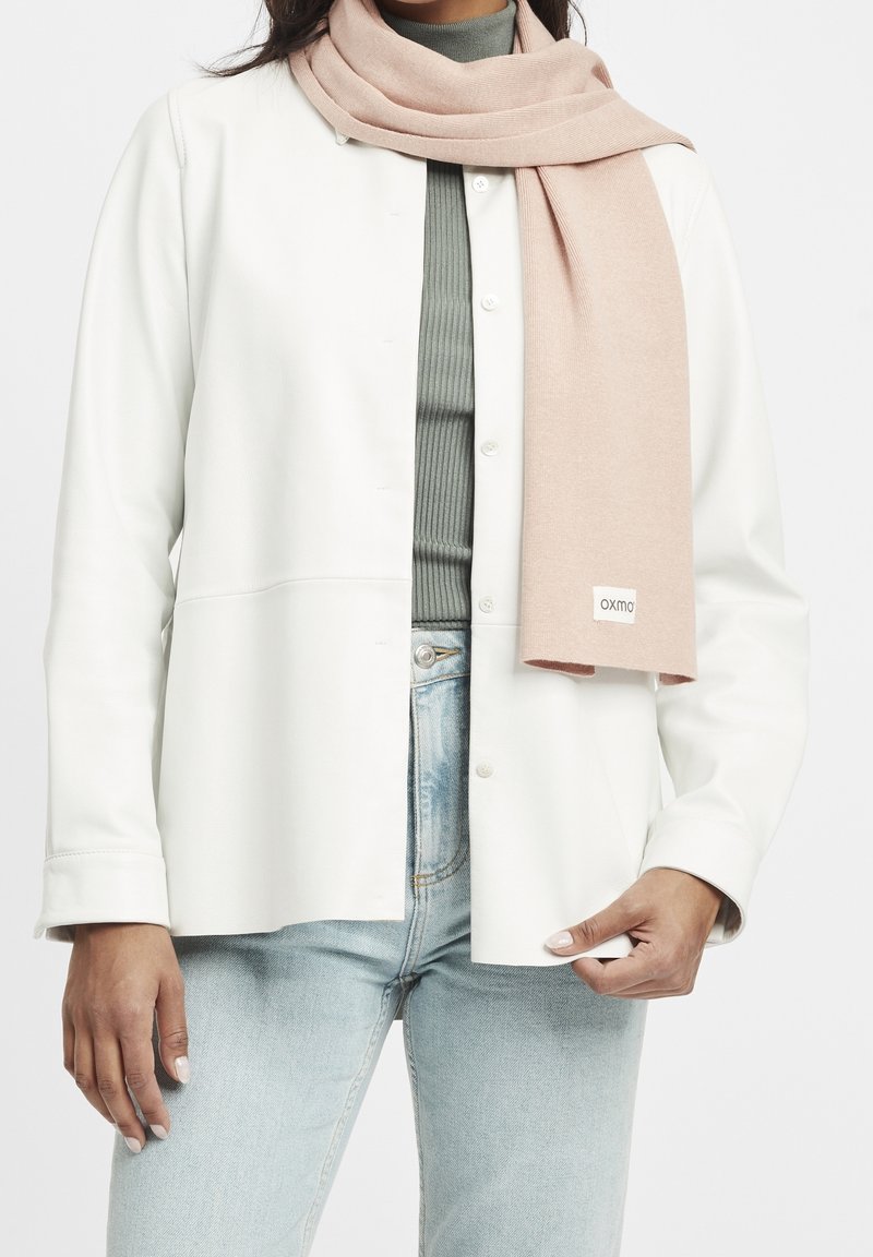 Personne portant une veste blanche boutonnée, un haut gris côtelé, un jean bleu clair et une écharpe rose pâle avec une petite étiquette à l'extrémité.