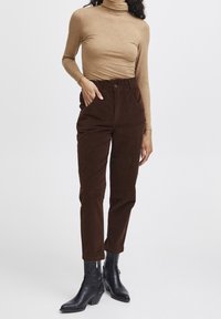 Kobieta w beżowym golfie, w brązowych spodniaczkach z sztruksu o wysokim stanie i czarnych botkach na obcasie sięgających poza łydki, stojąca z jedną ręką w kieszeni.