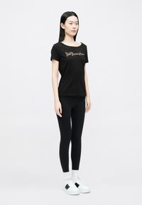 Fekete rövid ujjú póló széles nyakkivágással, aranyszínű felirattal. Fekete leggingssel és fekete részletekkel díszített fehér tornacipővel párosítva.