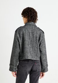 Klaus svart og grå fiskebensmønstret jakke med høy krage, knappede skuldre og brede mansjetter, kombinert med falmede svarte jeans.