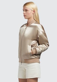 Saténová bomber bunda v metalické béžové barvě s pružnými manžetami a lemem; má přední zip a boční kapsy, spárovaná se krémovými šortkami.