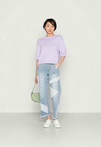 Sweater de malha canelada em lavanda, corte relaxado; jeans patchwork em azul claro com acentos brancos; bolsa verde; ténis brancos; de pé sobre um fundo neutro.