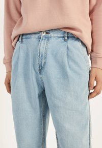 Pantalones de mezclilla azul claro con un corte relajado, frente plisado y cierre de botones. Presentan costuras sutiles y bolsillos laterales.