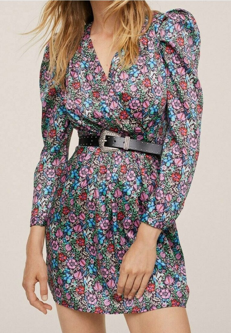 Femme portant une mini-robe à manches longues à motifs floraux avec des épaules bouffantes et une ceinture noire cloutée avec une boucle argentée.