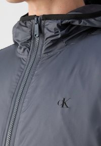 Grå zip-up jacka med hög krage och svarta detaljer. Har en logotyp på bröstet och är tillverkad av ett mjukt och lätt material.